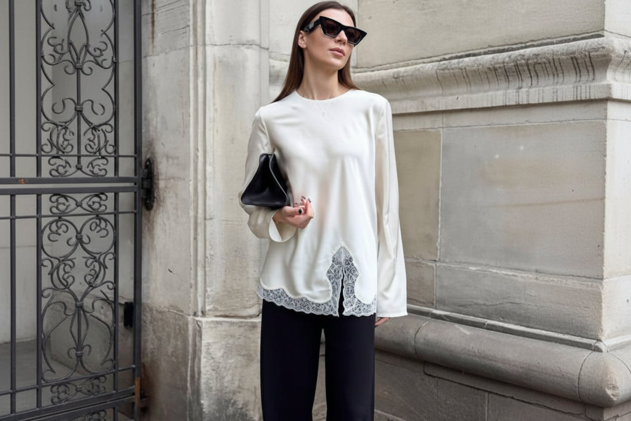 Woman wearing a white blouse with lace details and black pants, standing against a stone wall.
