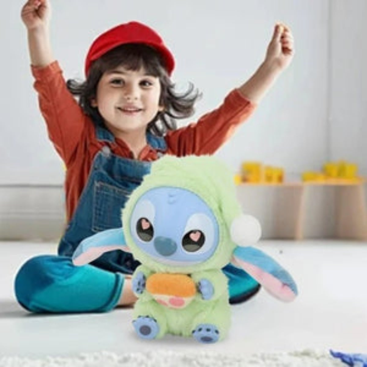 Child sitting on the floor with a large green plush toy resembling a cartoon character, wearing a red cap and blue overalls.