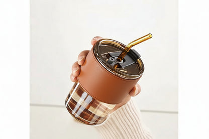 Hand holding a clear tumbler with a brown lid and straw, against a white background