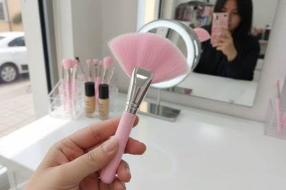 Hand holding a pink makeup brush with a blurred vanity setup in the background