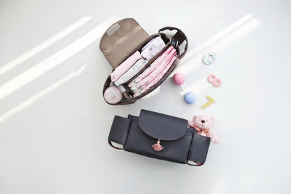 Black diaper bag with pink tassel on a white surface, containing baby items.