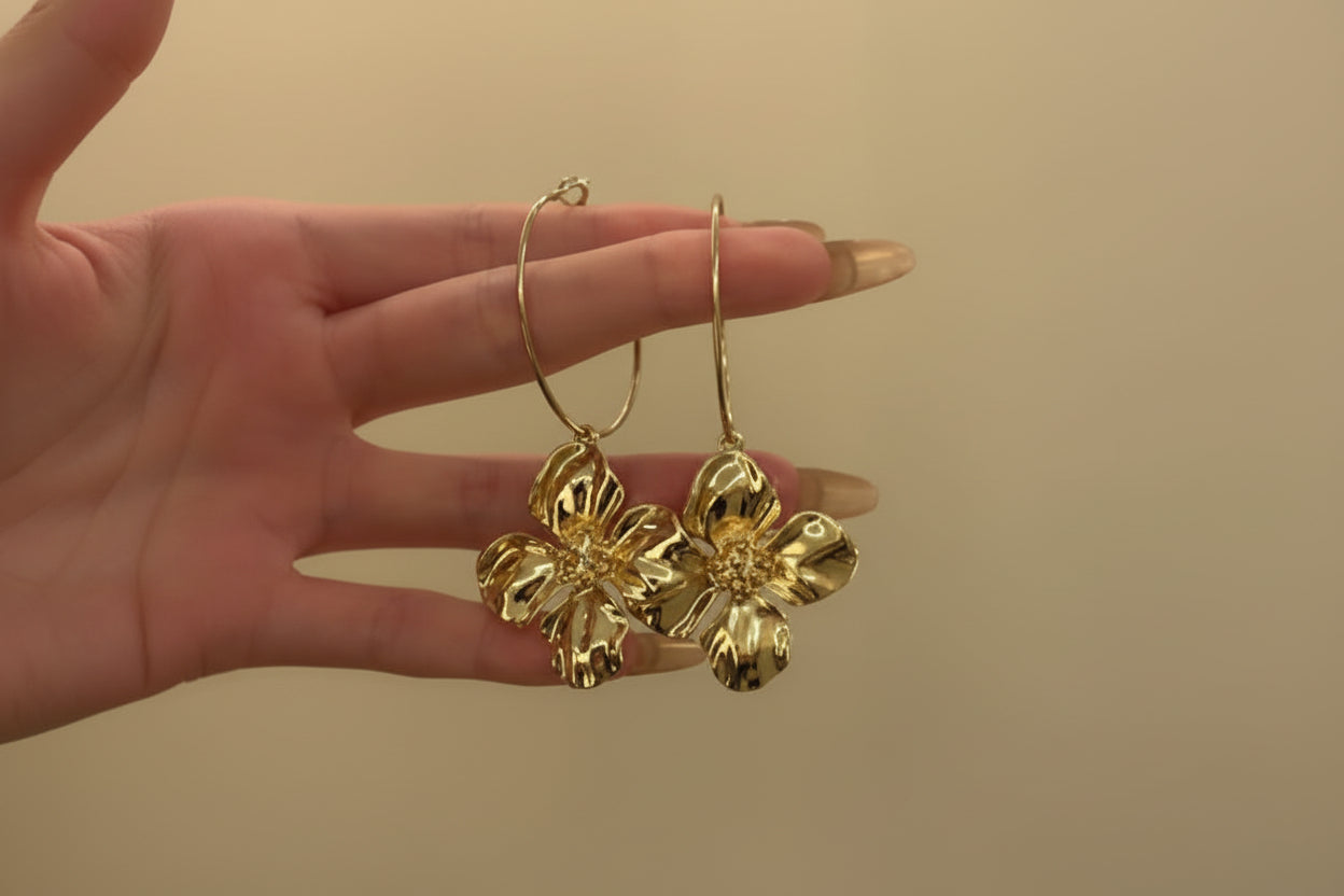 Gold flower-shaped earrings held by a hand against a beige background