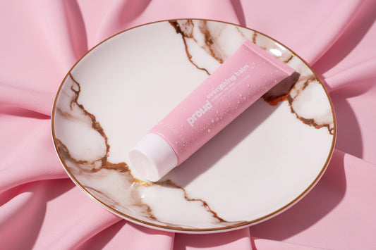 Pink tube of 'Proud Everything Balm' on a white background with coconut pieces.