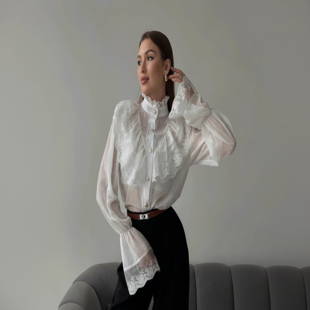 Woman wearing a white lace blouse and black pants sitting on a gray couch.
