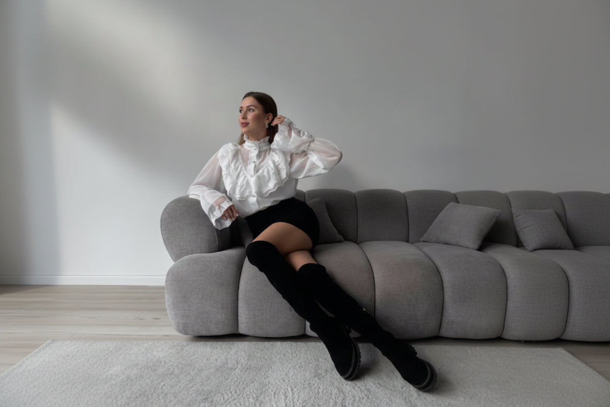 Woman sitting on a gray sofa wearing a white blouse and black knee-high boots in a minimalistic room.