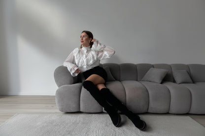 Woman sitting on a gray sofa wearing a white blouse and black knee-high boots in a minimalistic room.
