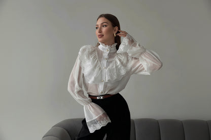 Woman wearing a white lace blouse and black pants sitting on a gray couch.