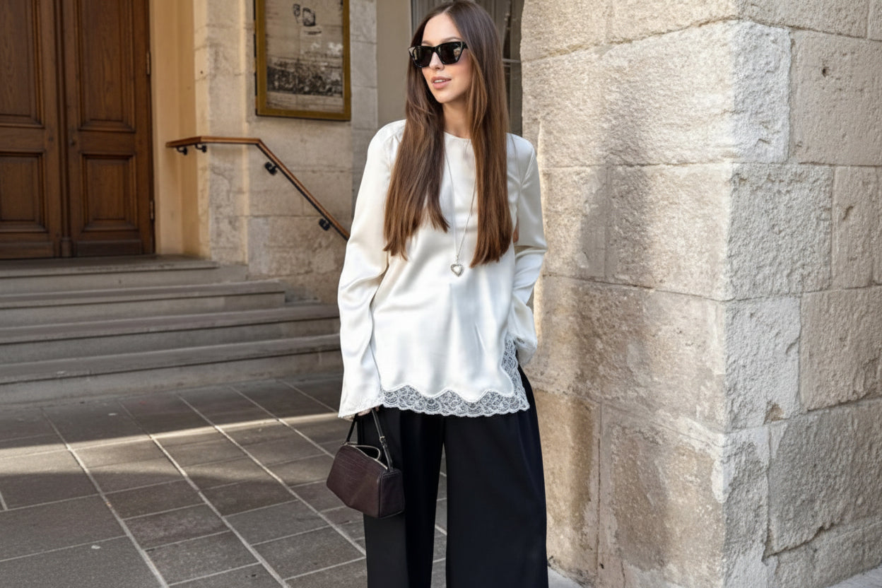 Woman wearing a white blouse with lace details and black pants, standing against a stone wall.