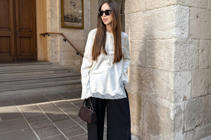 Woman wearing a white blouse with lace details and black pants, standing against a stone wall.