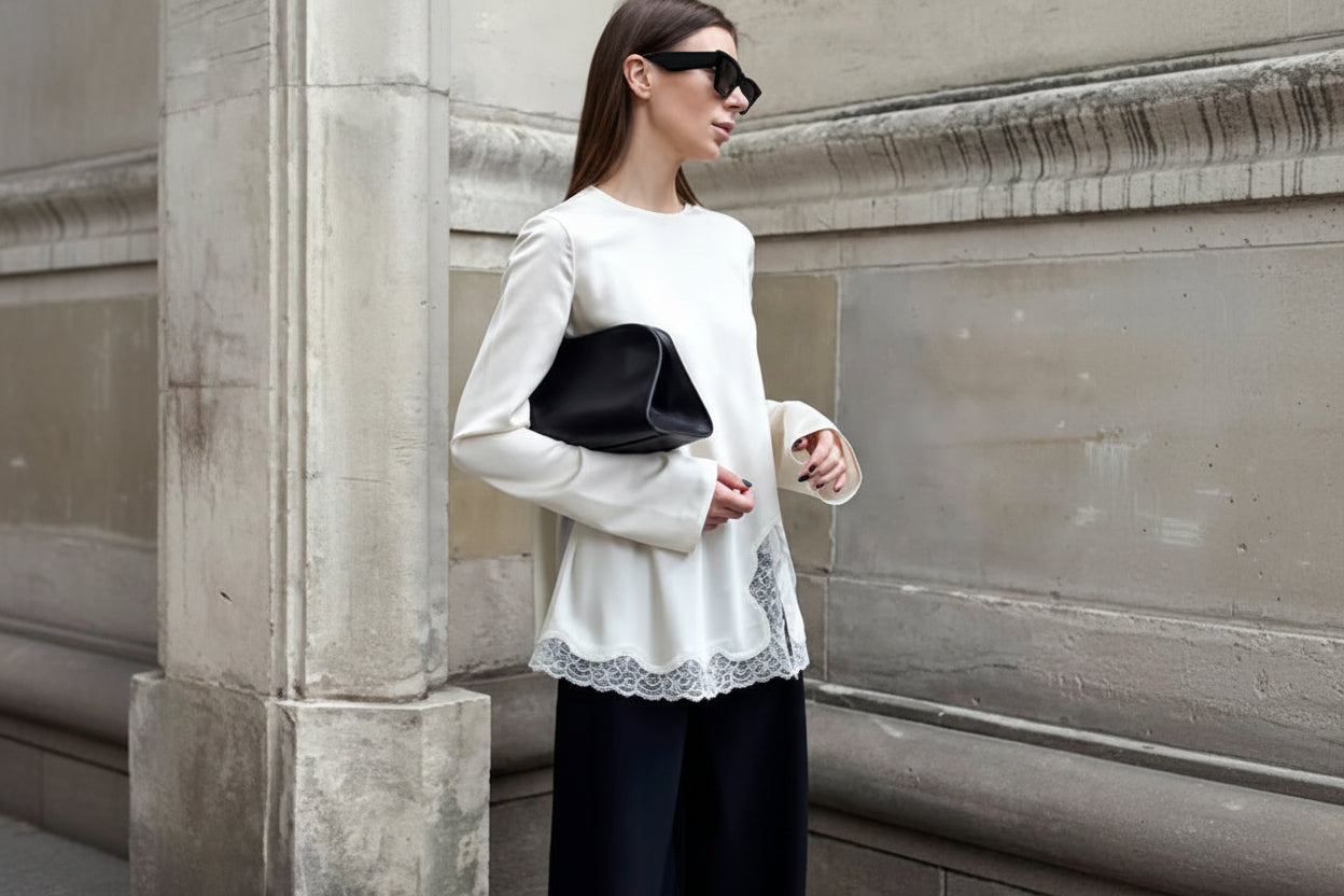 Woman in a white top with lace details and black pants standing against a stone wall.