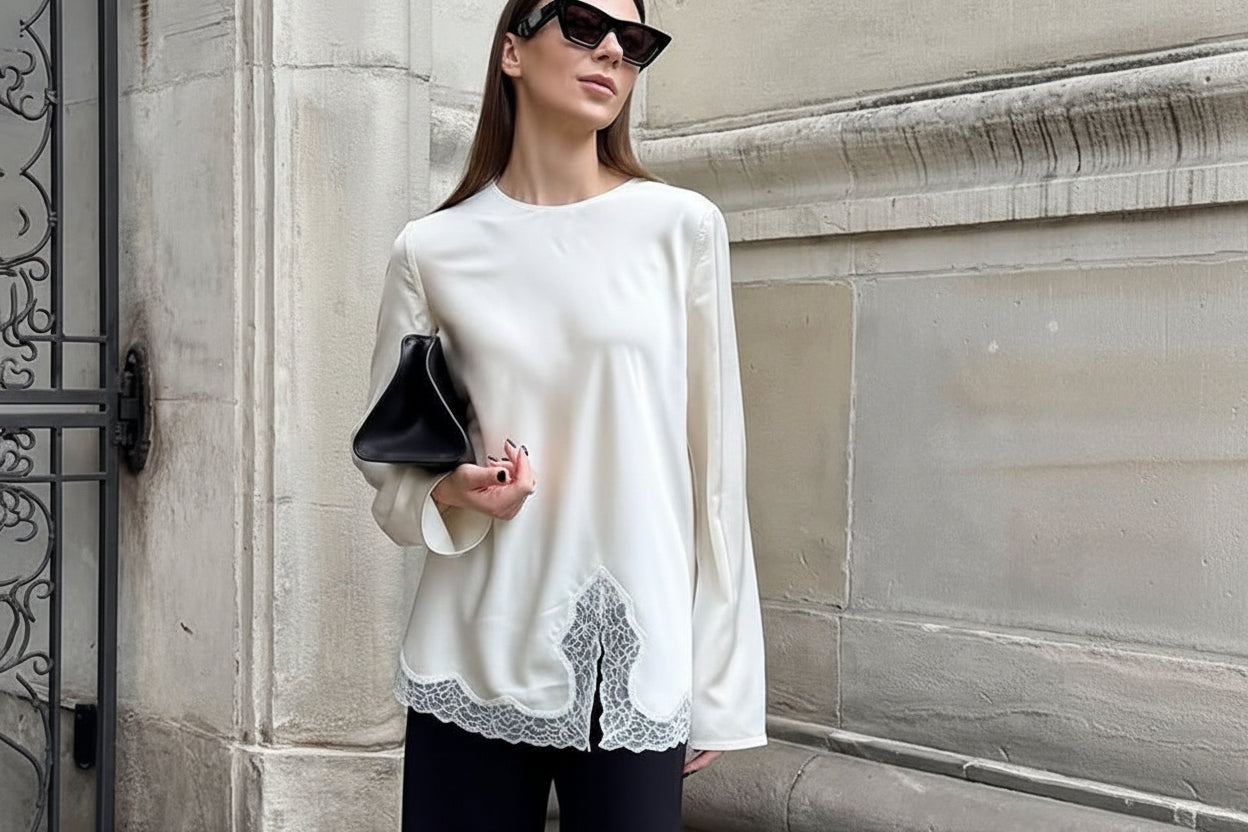Woman wearing a white blouse with lace details and black pants, standing against a stone wall.