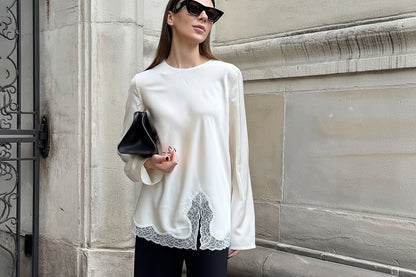 Woman wearing a white blouse with lace details and black pants, standing against a stone wall.