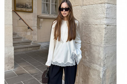Woman wearing a white blouse with lace details and black pants, standing against a stone wall.