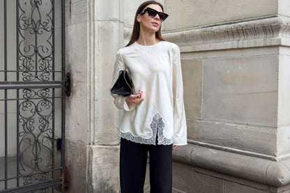 Woman wearing a white blouse with lace details and black pants, standing against a stone wall.