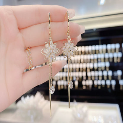 Gold flower-shaped earrings with dangling elements held by a hand against a blurred jewelry store background.