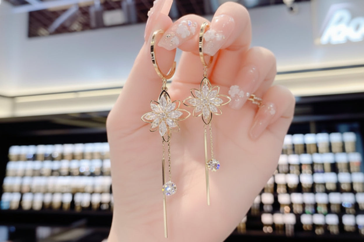 Gold flower earrings with dangling crystals held by a hand against a blurred background.