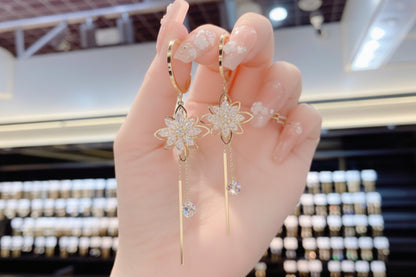 Gold flower earrings with dangling crystals held by a hand against a blurred background.