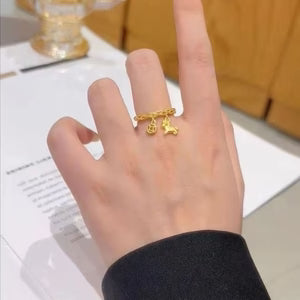 Gold ring with a small charm on a person's finger, set against a blurred indoor background.